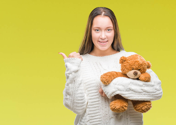 Young caucasian woman holding teddy bear over isolated background pointing and showing with thumb up to the side with happy face smiling - Photo, image