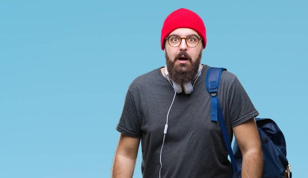 Νέοι hipster άνθρωπος φορώντας καπάκι κόκκινο μαλλί και σακίδιο πέρα από το απομονωμένο υπόβαθρο φοβάται και σοκαρισμένος με έκφραση έκπληξη, ο φόβος και ενθουσιασμένος πρόσωπο. - Φωτογραφία, εικόνα