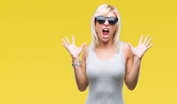 Young beautiful blonde woman wearing sunglasses over isolated background celebrating mad and crazy for success with arms raised and closed eyes screaming excited. Winner concept - Foto, Bild