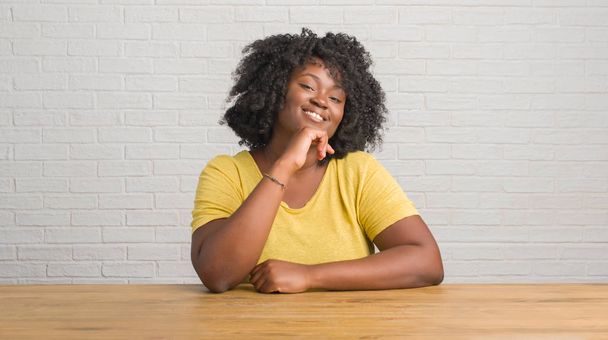 Mladá africká americká žena sedí na stole doma sebevědomým na kameru s úsměvem se zkříženýma rukama a ruku a na bradě. Pozitivní myšlení. - Fotografie, Obrázek