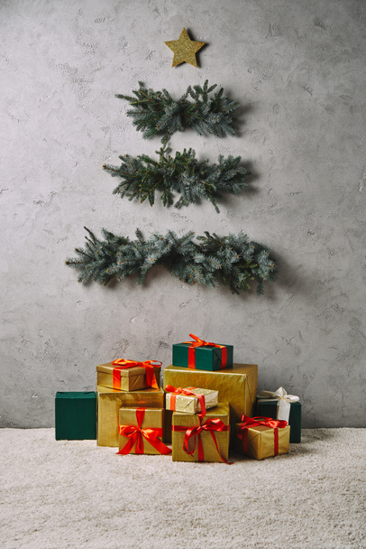 käsintehty joulukuusi roikkuu harmaalla seinällä, lahjapakkaukset matolla huoneessa
 - Valokuva, kuva
