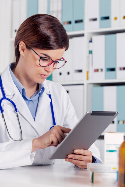 Jonge vriendelijke vrouw arts of verpleegkundige een stethoscoop met behulp van tablet pc in een kantoor dragen - Foto, afbeelding