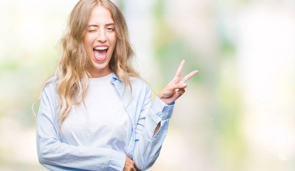 Beautiful young blonde woman over isolated background smiling with happy face winking at the camera doing victory sign. Number two. - Photo, Image