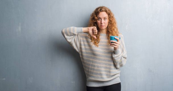 Молодая рыжая женщина через серую гранж-стену пьет чашку кофе с сердитым лицом, негативный знак, показывающий неприязнь с опущенными пальцами, концепция отказа
 - Фото, изображение