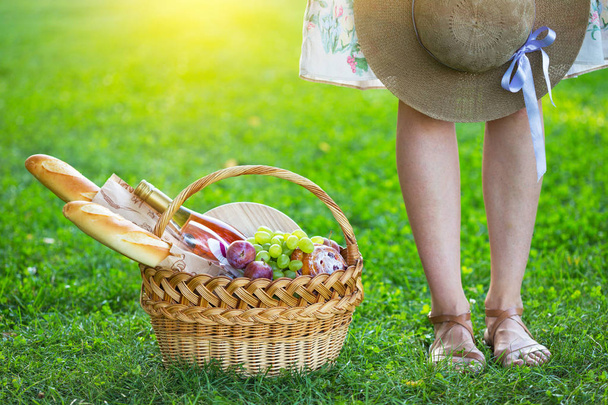 Лето - пикник на лужайке в парке. девушка и корзина для пикника с багетом, вином, бокалами, виноградом и рулоном
 - Фото, изображение