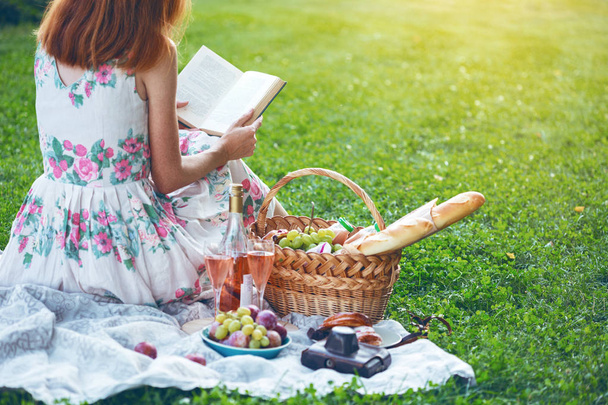 Summer - picnic in the meadow.  girl sitting reading a book and near a picnic basket and baguette, wine, glasses, grapes and roll - Foto, afbeelding