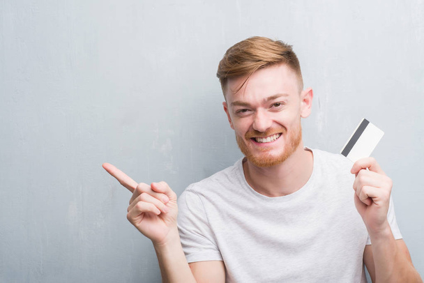 Jeune rousse homme sur le mur gris grunge tenant carte de crédit très heureux pointant avec la main et le doigt sur le côté
 - Photo, image