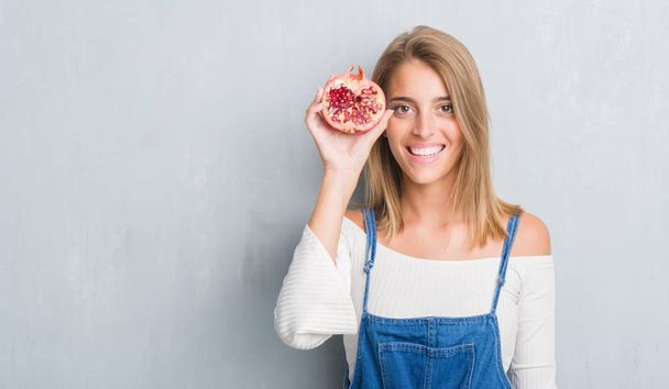 Beautiful young woman over grunge grey wall holding pomegranate with a happy face standing and smiling with a confident smile showing teeth - Photo, Image