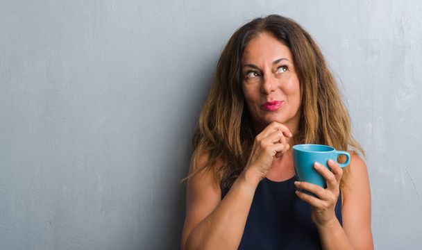 Mujer hispana de mediana edad de pie sobre la pared gris grunge beber taza de café cara seria pensando en la pregunta, idea muy confusa
 - Foto, imagen