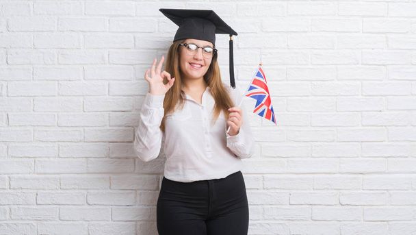 Mladé dospělé ženy bílá cihlová zeď nošení postgraduální víčko drží Velká Británie vlajka dělá ok cedulka s prsty, vynikající symbol - Fotografie, Obrázek