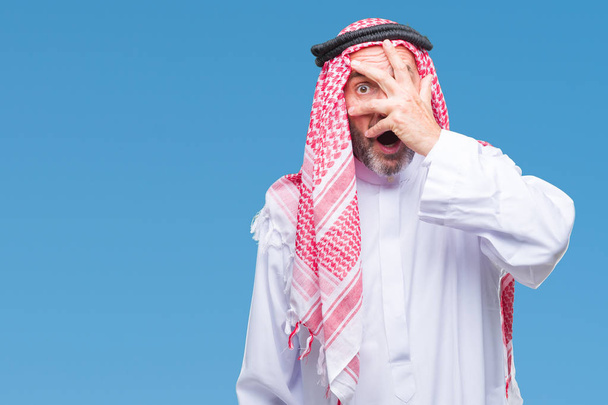 Älterer arabischer Mann mit Keffiyeh über isoliertem Hintergrund, der schockiert Gesicht und Augen mit der Hand bedeckt und mit verlegenem Gesichtsausdruck durch Finger blickt. - Foto, Bild