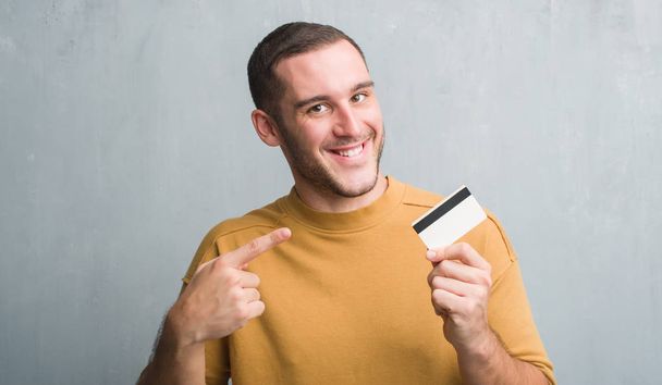 Młody człowiek kaukaski szary mur posiadania karty kredytowej bardzo szczęśliwy, wskazując z dłoni i palców - Zdjęcie, obraz
