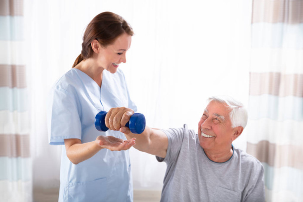 Young Female Nurse Helping Happy Senior Man With Dumbbell Exercise - Photo, image