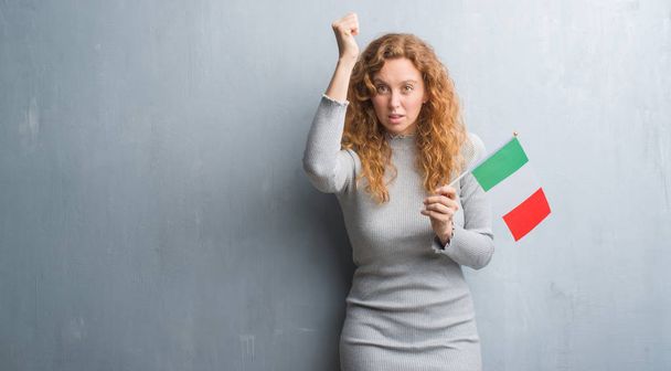Fiatal vöröshajú nő szürke grunge falon zászló Olaszország bosszús és csalódott kiabálás, a düh, őrült és kiabál, felemelt kézzel, harag koncepció - Fotó, kép