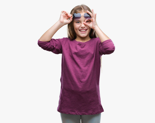 Young beautiful girl wearing sunglasses over isolated background doing ok gesture with hand smiling, eye looking through fingers with happy face. - Foto, Bild