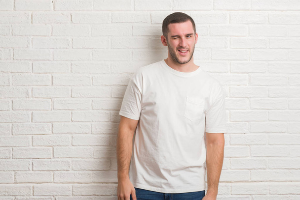 Young caucasian man standing over white brick wall winking looking at the camera with sexy expression, cheerful and happy face. - Photo, Image