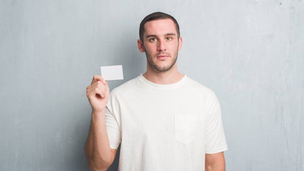 Молодий білий чоловік над сірою гранжевою стіною показує порожню візитну картку з впевненим виразом на розумному обличчі, думаючи серйозно
 - Фото, зображення
