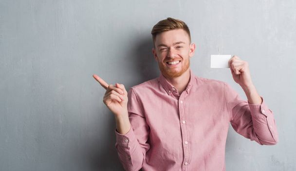 Jeune rousse homme sur le mur gris grunge tenant carte de visite vierge très heureux pointant avec la main et le doigt sur le côté
 - Photo, image