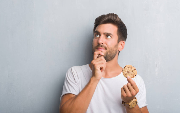 Handsome young man over grey grunge wall eating chocolate chip cooky serious face thinking about question, very confused idea - Photo, Image