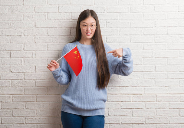 Fiatal kínai nő gazdaság zászlaja Kína meglepetés arc mutatóujjal magát tégla falon - Fotó, kép