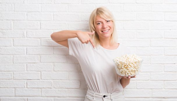 Adult caucasian woman over white brick wall eating pop corn with surprise face pointing finger to himself - Фото, зображення