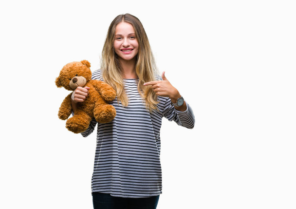 Young beautiful blonde woman holding teddy bear plush over isolated background with surprise face pointing finger to himself - Фото, зображення