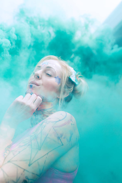 Wide portrait of blonde caucasian woman with tattoos using green smoke bomb in an urban location - Photo, Image