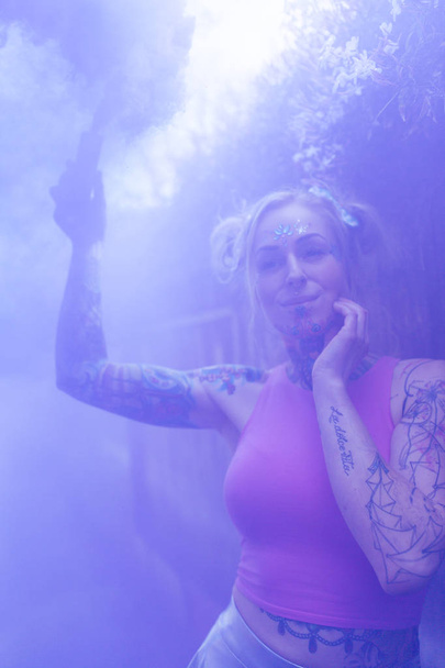Amplo retrato de mulher loira caucasiana com tatuagens usando bomba de fumaça azul em um local urbano
 - Foto, Imagem
