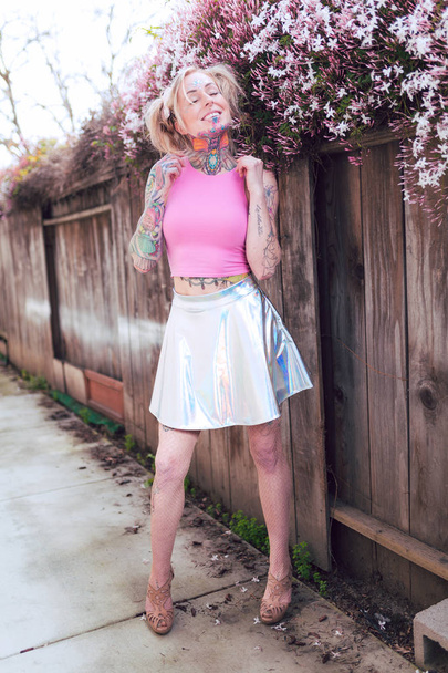 Wide portrait of blonde caucasian woman with tattoos near pink flowers wearing heels and shiny skirt - Foto, Bild