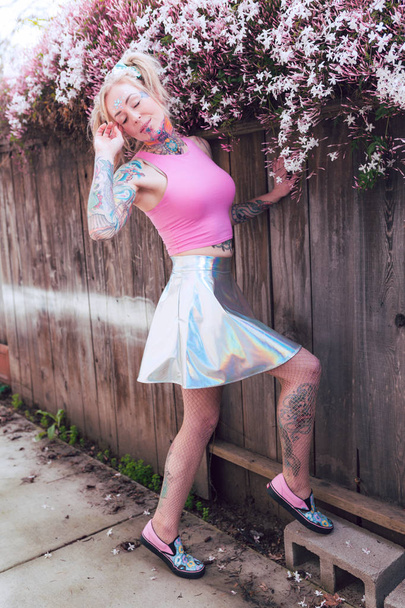 Wide portrait of blonde caucasian woman with tattoos near pink flowers wearing heels and shiny skirt - Photo, image