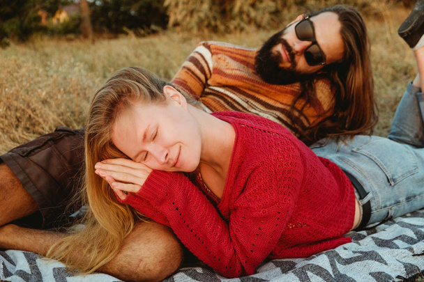 Een hipster paar blonde vrouw en brunette man met lange haren is koelen en knuffelen op de deken in de gouden zonsopgang. - Foto, afbeelding