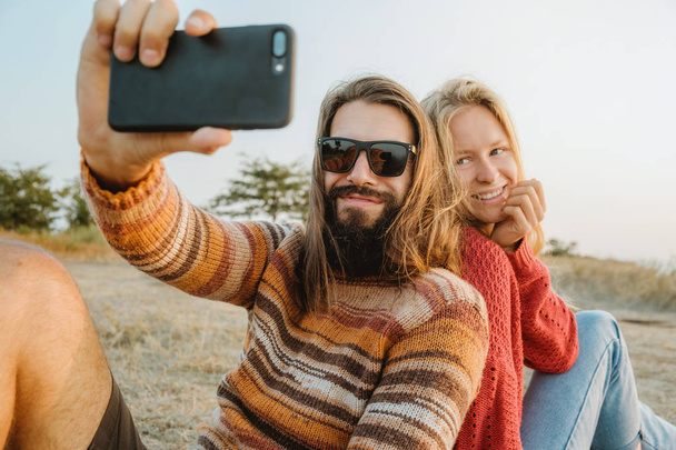 Una coppia hipster di donna bionda e uomo bruna con i capelli lunghi che indossano maglioni e fanno selfie all'aperto
. - Foto, immagini