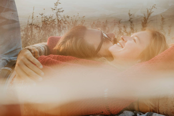 Una coppia hipster di donna bionda e uomo bruna con i capelli lunghi in maglioni che si rilassano e si abbracciano sulla coperta all'aperto all'alba
. - Foto, immagini
