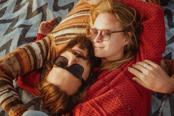 A hipster couple of blond woman and brunette man with long hair in sweaters chilling and hugging on blanket outdoors in sunrise. - Φωτογραφία, εικόνα
