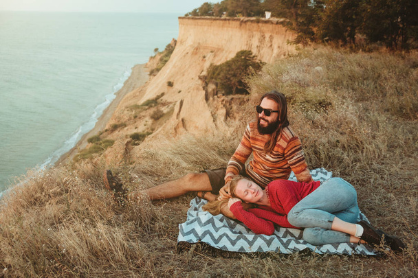 A hipster couple of blond woman and brunette man with long hair is chilling and hugging on blanket on rocky coast with seaview - Photo, Image