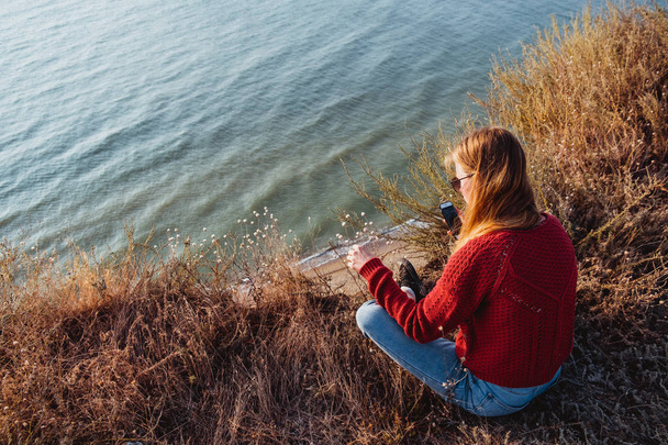 Une jeune photographe rousse en pull rouge prenant des photos de fleurs sur une côte rocheuse
. - Photo, image