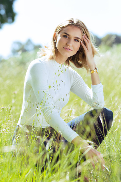 Attractive young blonde Caucasian woman in tight white top and denim posing outside in long grass - Фото, изображение