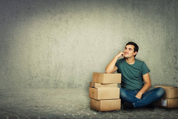 Pensive young man sitting and leaning on cardboard boxes hand on cheek and dream. Ready for moving to a new house over grey wall background. - Photo, Image