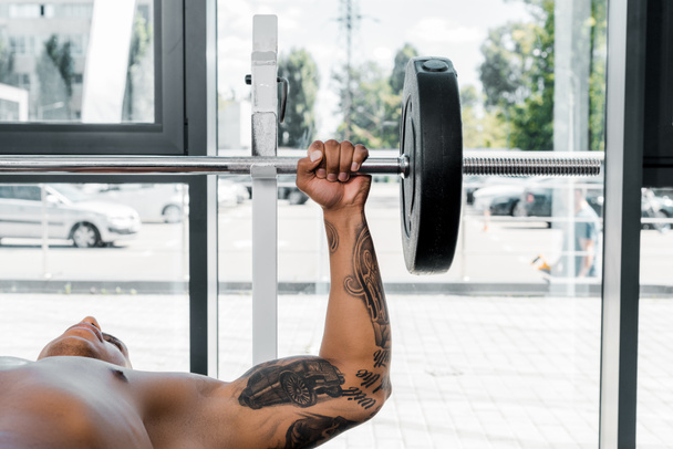muscular tattooed african american sportsman lying on bench and lifting barbell in gym - Photo, Image