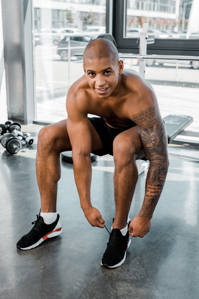 young muscular african american sportsman tying shoelaces and smiling at camera in gym - Zdjęcie, obraz