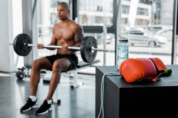 close-up view of boxing glove, skipping rope and bottle of water and sportsman lifting barbell behind in gym - Foto, imagen