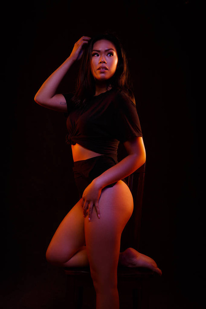 Fit young asian woman in black one piece poses on a chair under colored lights - Foto, Imagen
