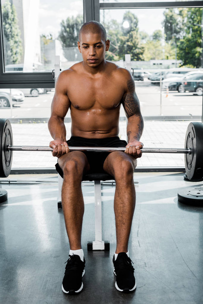 athletic young african american sportsman sitting on bench and lifting barbell in gym - Fotó, kép