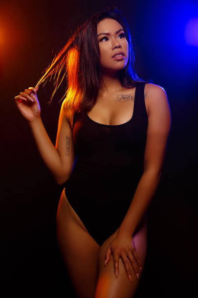 Fit young asian woman in black one piece poses against black under  orange and blue colored lights - Photo, Image