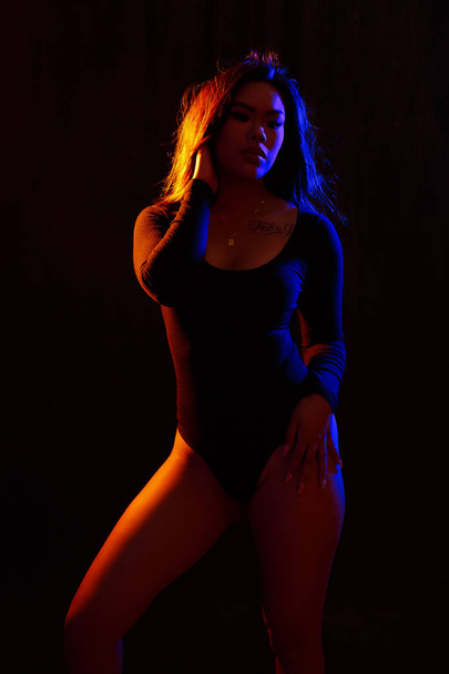 Fit young asian woman in black one piece and necklace poses against black under  orange and blue colored lights - Foto, Imagem