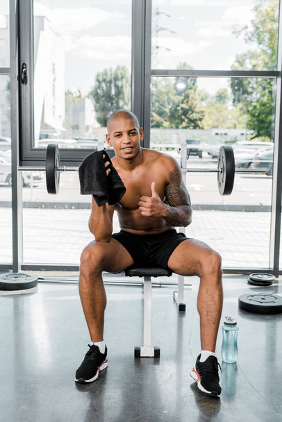 muscular young african american sportsman with towel showing thumb up and smiling at camera in gym - 写真・画像