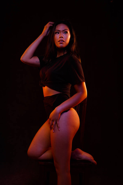 Fit young asian woman in black one piece poses on a chair under colored lights - 写真・画像