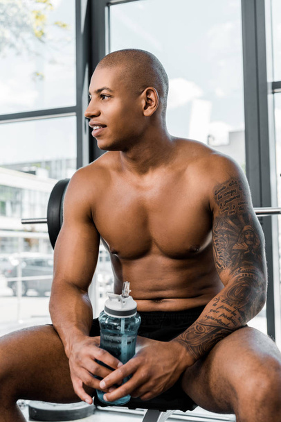 handsome smiling young sportsman holding bottle of water and looking away while sitting in gym - Foto, Bild