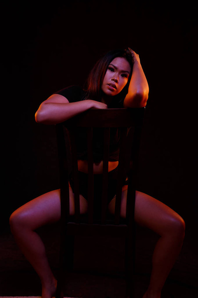 fit junge asiatische Frau in schwarz Einteiler posiert auf einem Stuhl unter farbigen Lichtern - Foto, Bild