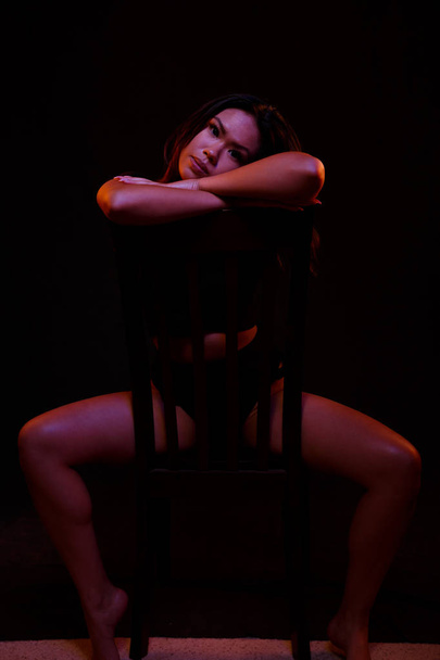 Fit young asian woman in black one piece poses on a chair under colored lights - Фото, изображение